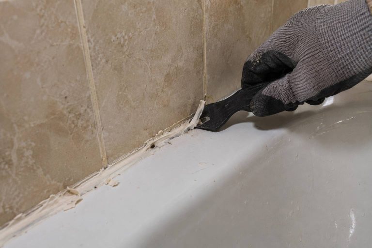 Une main gantée utilise un grattoir pour retirer l'ancien mastic du joint entre un mur carrelé et une baignoire.